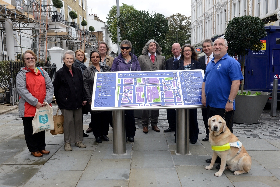 Image depicts a group of visually impaired people, volunteers and staff standing around the Exhibition road tactile map for a group photograph. They are all smiling. There is also a guide dog on the right of the picture. The dog is looking towards the camera.