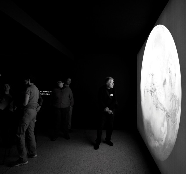 This image depicts VI's at the entrance to the Moving to Mars exhibition, with a large image of Mars illuminated on the right wall. The photograph is a black and white image, with the left side very dark.